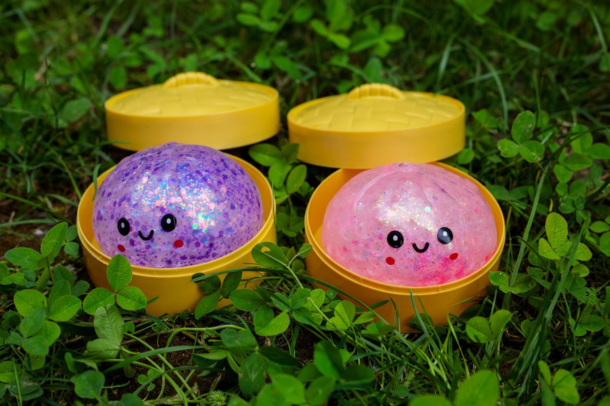 Two kawaii glitter squishy dumpling toys, one purple and one pink, in yellow steamer containers on grass